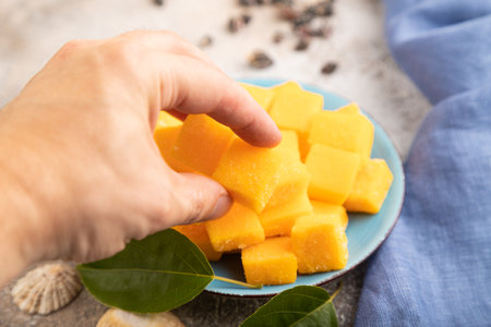 Dried and candied mango cubes on blue plate with hand on brown concrete background and linen textile. Side view, close up, selective focus, vegan, vegetarian food concept.の写真素材