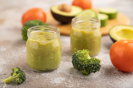 Baby puree with vegetable mix, broccoli, tomatoes, cucumber, avocado infant formula in glass jar on brown concrete background. Side view, close up, selective focus, artificial feeding concept.の写真素材
