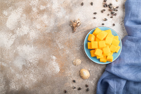 Dried and candied mango cubes on blue plate on brown concrete background and linen textile. Top view, flat lay, copy space, vegan, vegetarian food concept.の写真素材