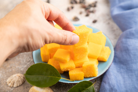 Dried and candied mango cubes on blue plate with hand on brown concrete background and linen textile. Side view, close up, selective focus, vegan, vegetarian food concept.の写真素材