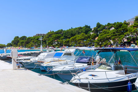 7 July 2021 Brela, Croatia: Many private yachts are parked near the pier. Tourists rest on a pine...