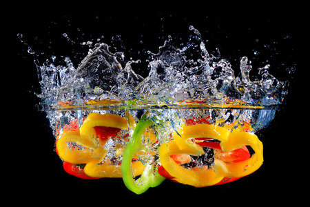 Red yellow and green pepper slices fall into water, on black backgroundの写真素材