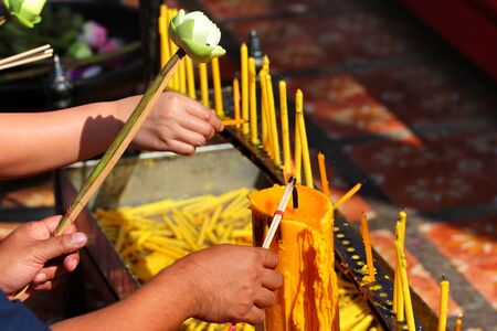 Hands holding incense sticks, lighting from the burning yellow candle. Traditional Thailand buddhist concept.の写真素材