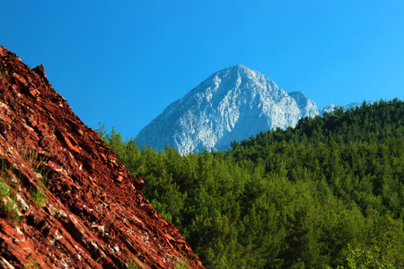 Mountain peak against blue sky behind green forest and red hillside