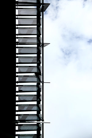 Underside view of metal staircase with mesh treads silhouetted against cloudy skyの写真素材