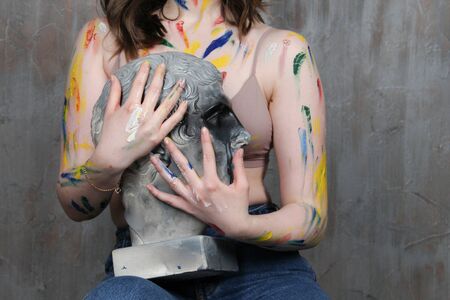 A young woman who has gotten dirty with paint holds a sculpture of a manâs head with a Greek profile. Art, creativity, hobby concept, drawing process during the pandemic self-isolation COVID-19. stay home, 2019 nKoV preventionの写真素材