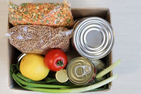 cardboard box with food supplies crisis food stock for quarantine isolation period on light background. Canned food, buckwheat, legumes, lemon, tomato, green onion. Food delivery, Donation, coronavirus quarantine.の写真素材