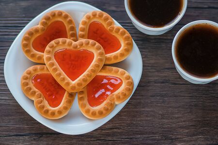 Heart-shaped cookies with strawberry jam on a white plate and two white cups of coffee on a brown wooden background. Love concept. View from above. Soft focus.の写真素材