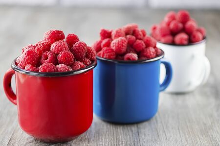 Ripe organic raspberries in blue, red and white metal mugs on gray background. Soft focus.の写真素材
