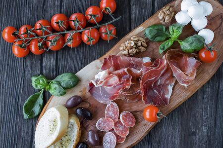 Prosciutto, bread, olives, walnut, mozzarella, salami, basil and cherry tomatoes on  brown wooden board.  Mediterranean kitchen. Soft focus.の写真素材