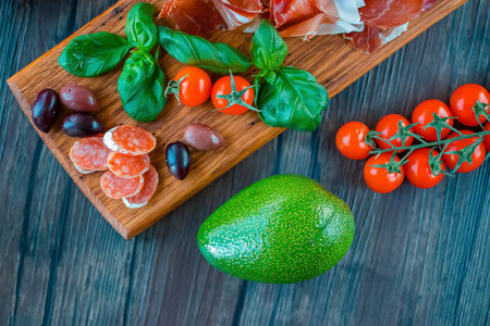 Prosciutto, olives, salami, basil, avocado and cherry tomatoes on  brown wooden board. ...