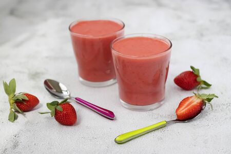 Organic strawberry smoothies in glass cups, bright spoons and berries of ripe strawberries on a gray cement background. Soft focus.の写真素材
