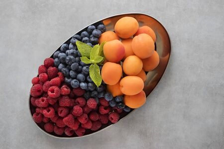 Ripe organic raspberries, blueberries and apricots in a metal oval plate on a gray background. Top view. Soft focus.の写真素材