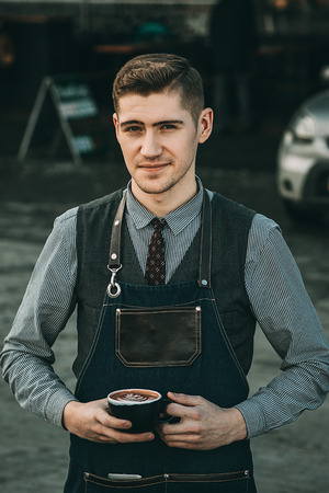 Cropped image of handsome barista in apron holding a cup of coffee and smiling while working at the bar counter in cafeの写真素材