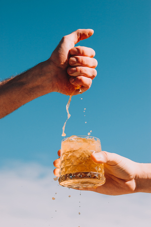 Squeeze out juice from orange by hands into lemonade with ice. Cloud sky background. Summer drink conceptの写真素材