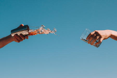 Hard spilled ice summer drink from shaker to glass. Hands of bartender. Blue sky background. Summer drink concept.の写真素材