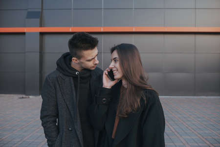 Young loving couple with coffee on black industrial background looks into boyfriend phoneの写真素材