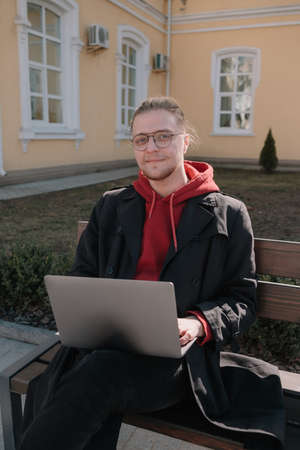 Young man university student online learning via laptop computer, sitting on a bench on a campus. Stylish hipster guy experienced freelancer working distance on notebook while restingの写真素材