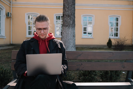 Young man university student online learning via laptop computer, sitting on a bench on a campus. Stylish hipster guy experienced freelancer working distance on notebook while restingの写真素材