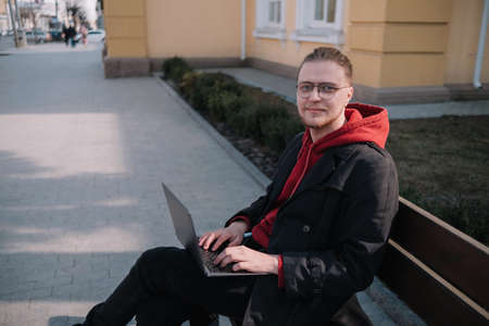 Young man university student online learning via laptop computer, sitting on a bench on a campus. Stylish hipster guy experienced freelancer working distance on notebook while restingの写真素材