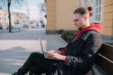 Young man university student online learning via laptop computer, sitting on a bench on a campus. Stylish hipster guy experienced freelancer working distance on notebook while restingの写真素材