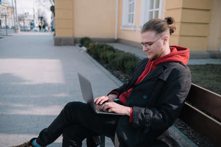 Young man university student online learning via laptop computer, sitting on a bench on a campus. Stylish hipster guy experienced freelancer working distance on notebook while restingの写真素材
