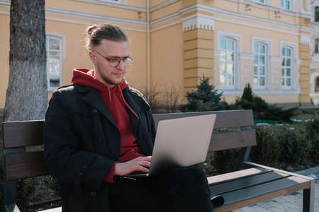 Young man university student online learning via laptop computer, sitting on a bench on a campus. Stylish hipster guy experienced freelancer working distance on notebook while restingの写真素材