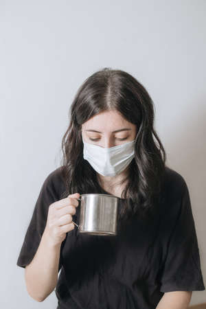 Young woman with a cup of tea at the window during quarantine wearing face mask worried about the new lifestyle.の写真素材