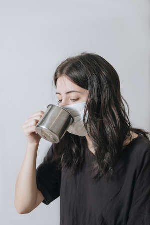 Young woman with a cup of tea at the window during quarantine wearing face mask worried about the new lifestyle.の写真素材