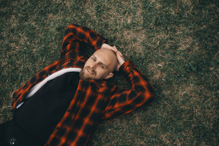 Young bald man with a beard lying on the grass in the park and smiling looking at the camera