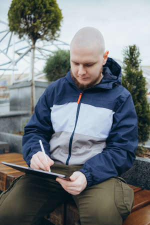 From below of hipster man using modern tablet with stylus while creating new project and sitting on street near glass skyscraperの写真素材