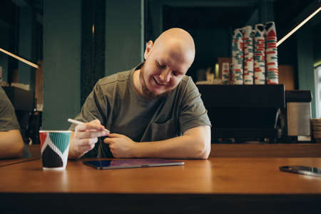 Young creative web designer in casualwear having coffee with cookies and drawing with stylus on tablet while working over new websiteの写真素材