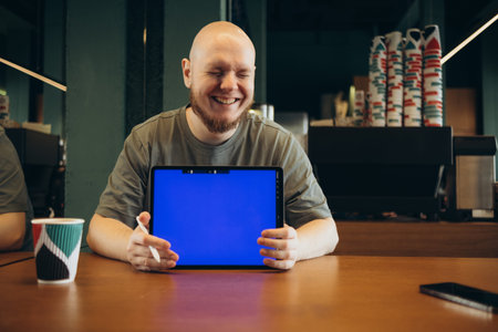 Cheerful male pointed at empty blue screen of tablet by finger while sitting on bench near modern...
