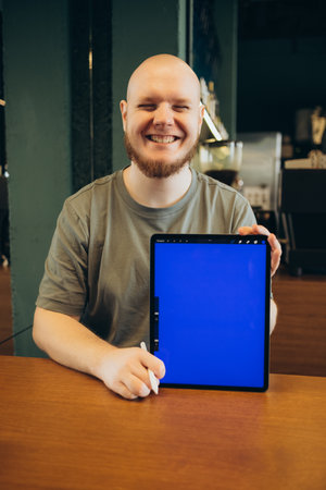 Cheerful male pointed at empty blue screen of tablet by finger while sitting on bench near modern cafe and looking at cameraの写真素材