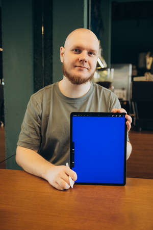 Cheerful male pointed at empty blue screen of tablet by finger while sitting on bench near modern cafe and looking at cameraの写真素材