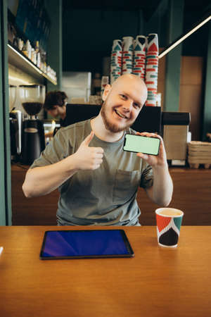 Cheerful male pointed at empty blue screen of tablet by finger while sitting on bench near modern cafe and looking at cameraの写真素材