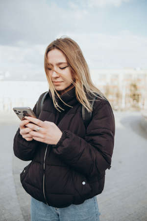gorgeous beautiful young woman with blonde hair messaging on the smart-phone at the city street background. pretty girl having smart phone conversation in sun flare.の写真素材
