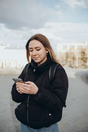 gorgeous beautiful young woman with blonde hair messaging on the smart-phone at the city street background. pretty girl having smart phone conversation in sun flare.の写真素材