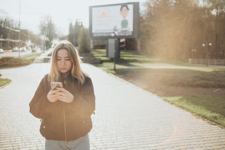 gorgeous beautiful young woman with blonde hair messaging on the smart-phone at the city street background. pretty girl having smart phone conversation in sun flare.の写真素材