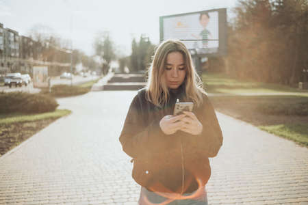 gorgeous beautiful young woman with blonde hair messaging on the smart-phone at the city street background. pretty girl having smart phone conversation in sun flare.の写真素材