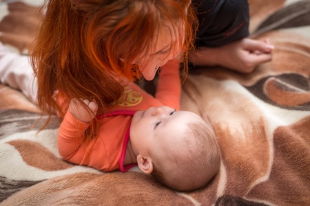 little baby lying on the bed playing with a red-haired mother and smiling happilyの写真素材