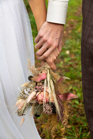 The bride and groom are holding hands. In hands a bouquet of dried flowers in boho styleの写真素材