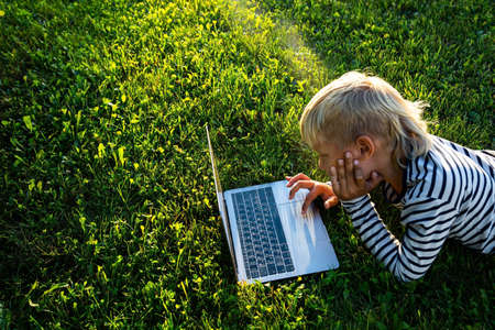 The boy is engaged in the Internet outdoors. The child lies on the grass with a laptop.の写真素材