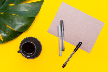Blank sheet of paper, felt-tip pens, monstera leaf and a cup of black coffee on a yellow background....
