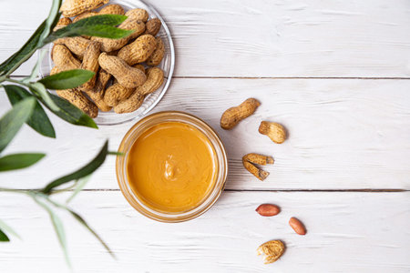 A jar of peanut butter with a bunch of peanuts on a white wooden table with an olive branch. Homemade peanut butter, natural, healthy food. The modern wellness and vegan concept.の写真素材