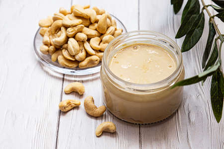 A jar of cashew butter with a bunch of fresh cashew on a white wooden table with an olive branch. Homemade cashew butter, natural, healthy food. The modern wellness and vegan concept.の写真素材