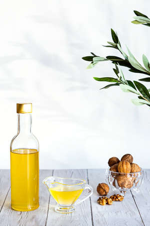 Walnut oil and a bunch of nuts on a white wooden table. Modern still life with oil bottle and glass bowl with walnuts, olive branch and copy space.の写真素材
