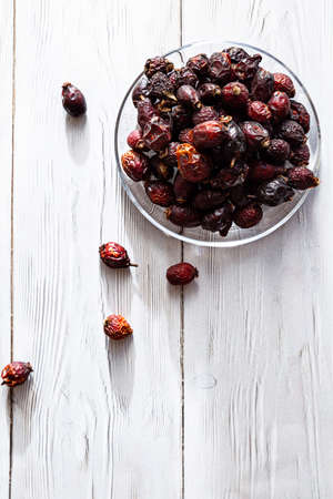 Dried rose hip berries in a glass plate on a white wooden table. Concept of traditional medicine.Organic rose hip - Antioxidant, source of vitamin C, immunity protection. Superfoods.の写真素材