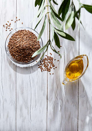 Linseed oil and a bunch of flax seeds on a white wooden table. Modern still life glass ball with oil and linseed, olive branch and copy space.の写真素材