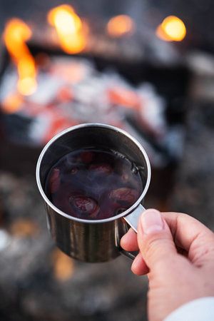 Woman hand hold rosehip tea in steel mug against bonfire. Eco-friendly tourism, sustainable...